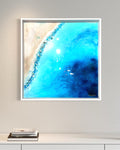A framed abstract painting depicting a stylized beach scene with blue and green colors, crystals on canvas, set in a white oak frame.